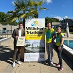 Stadtwerke-Geschäftsführer Tafil Pufja übergibt im Freizeitbad de Bütt die gesponserten Starterbeutel an Johannes Hüsch und Stephanie Grund-Liethen vom TV Schiefbahn. Im Hintergrund: Triathlon-Banner und Palmen.