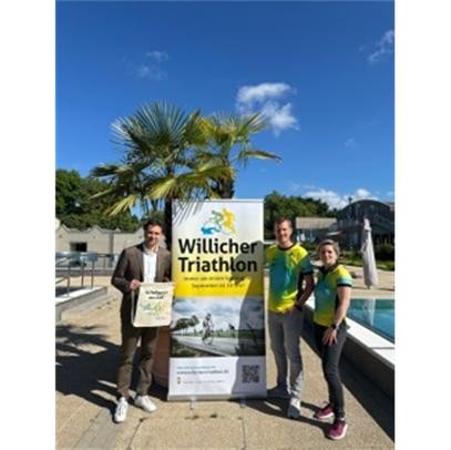 Stadtwerke-Geschäftsführer Tafil Pufja übergibt im Freizeitbad de Bütt die gesponserten Starterbeutel an Johannes Hüsch und Stephanie Grund-Liethen vom TV Schiefbahn. Im Hintergrund: Triathlon-Banner und Palmen.