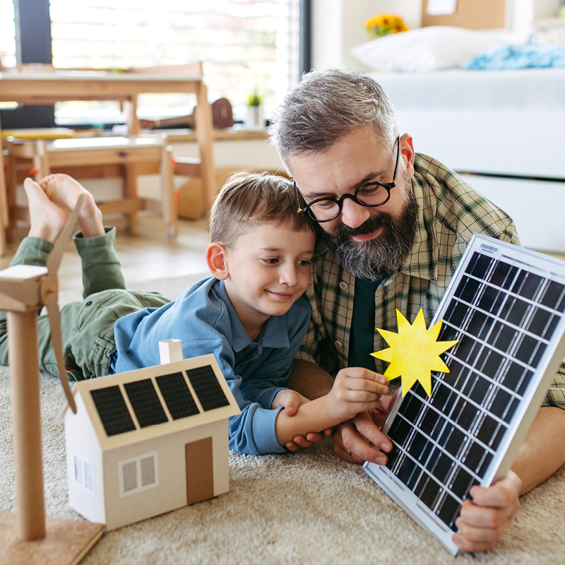 Ein Vater und sein Sohn liegen auf dem Teppich und betrachten spielerisch ein Solarmodell mit Sonnensymbol – Symbolbild für den bewussten Umgang mit Ökostrom und erneuerbarer Energie im Alltag