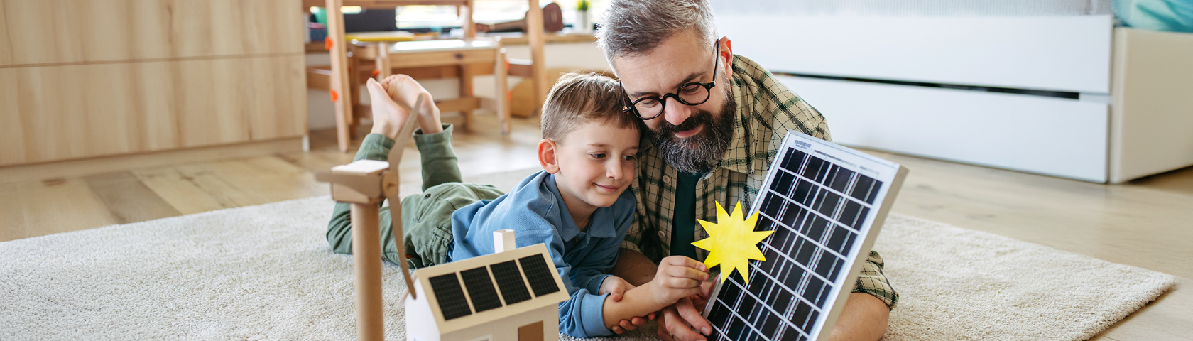 Ein Vater und sein Sohn liegen auf dem Teppich und betrachten spielerisch ein Solarmodell mit Sonnensymbol – Symbolbild für den bewussten Umgang mit Ökostrom und erneuerbarer Energie im Alltag