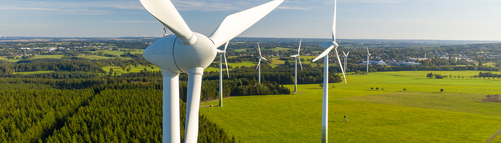 Blick auf mehrere Windräder in einer grünen Landschaft mit Stromtrassen im Hintergrund – Symbolbild für die regionale Stromnetzinfrastruktur