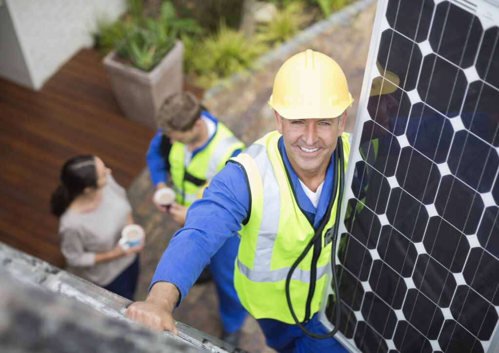 Handwerker montiert Solarmodul auf Dach, unten sprechen zwei Personen mit Kaffee in der Hand