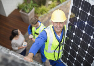 Handwerker montiert Solarmodul auf Dach, unten sprechen zwei Personen mit Kaffee in der Hand