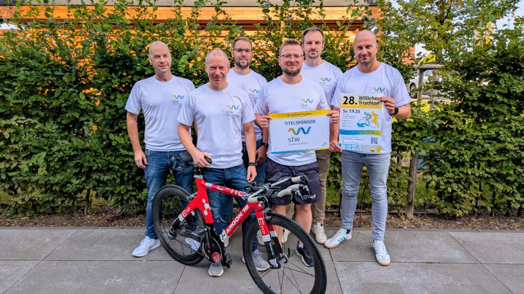 Stadtwerke-Team mit sechs Männern und Rennrad posiert vor einer Hecke, zwei halten Plakate zum Willicher Triathlon