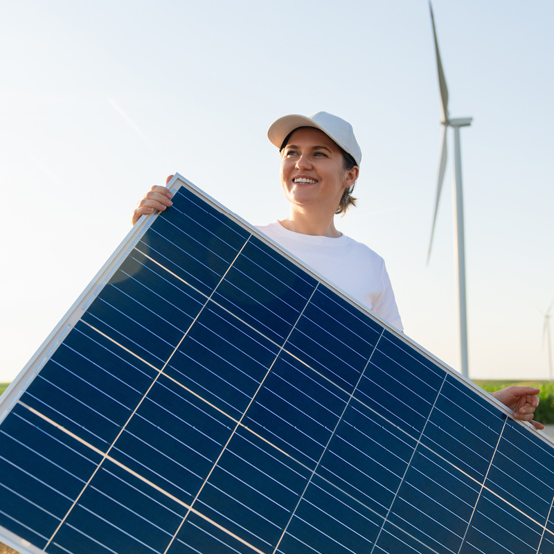 Frau trägt Solarmodul auf Feld mit Windrädern im Hintergrund