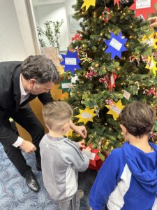 Erwachsener spricht mit Kind am Weihnachtswunschbaum mit bunten Wunschsternen.