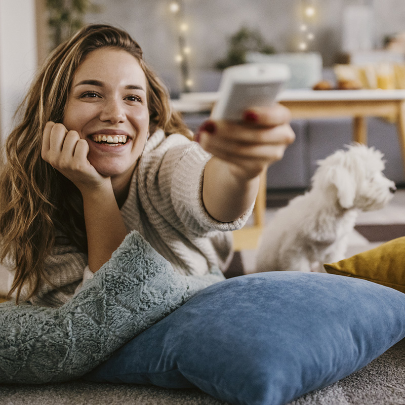 Frau liegt entspannt auf dem Boden mit Fernbedienung, Hund sitzt im Hintergrund