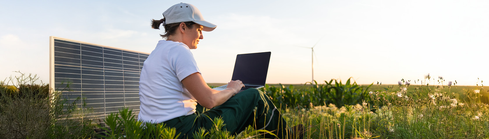 Kundin sitzt mit Laptop vor Photovoltaikanlage auf Feld bei Sonnenuntergang